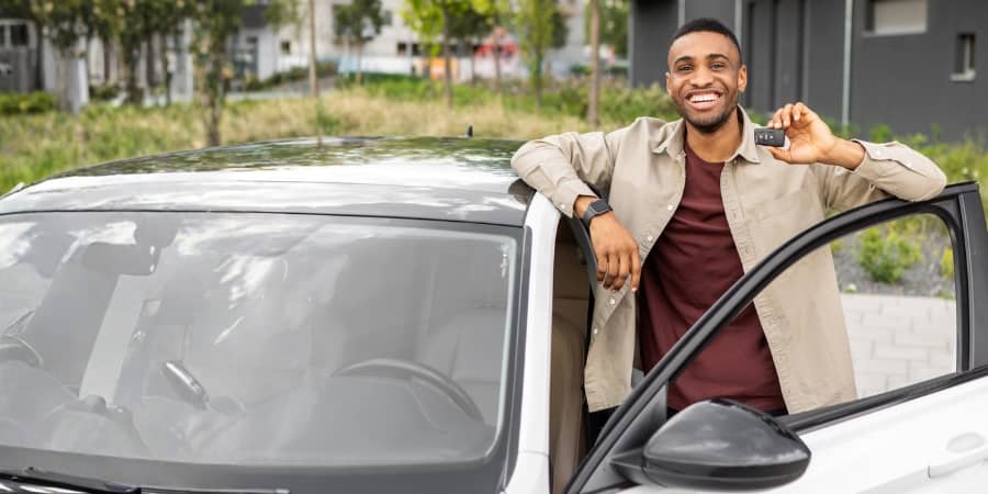 A young male adult standing next to the driver-side of a car holding keys and smiling.