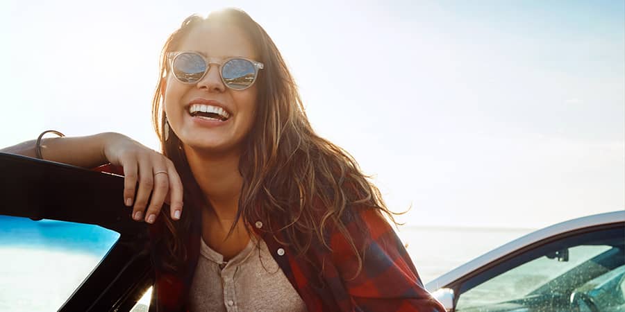 A young, smiling woman standing outside a car wearing glasses.