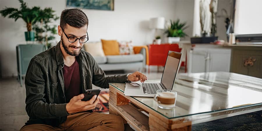 A middle-aged man sitting at a table using a laptop and smart phone.