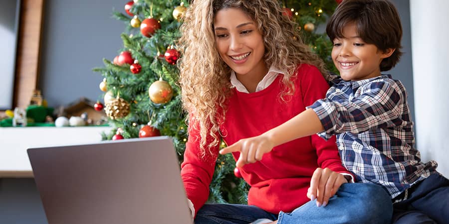 A young mother and her son sitting in front of a christmas tree while using a laptop.