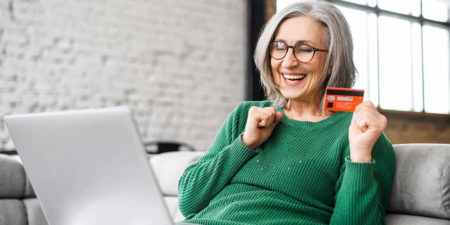 An elderly woman sitting on a couch with a laptop open and a card in hand.