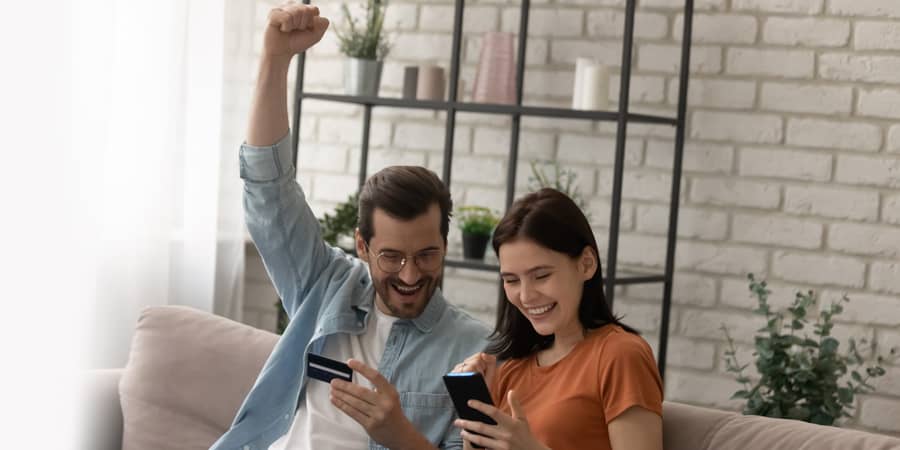 A young couple sitting on a couch and celebrating while looking at a credit card and mobile phone.