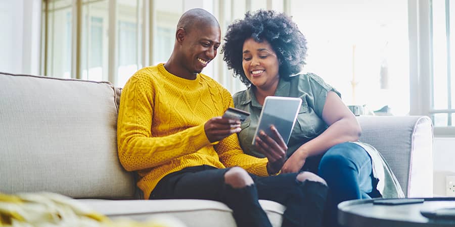 A young couple sitting on a couch with one holding a tablet and a credit card.