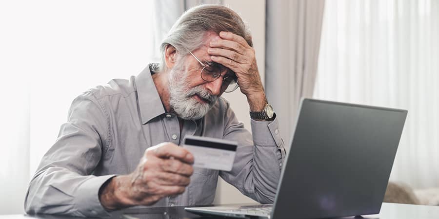 A distressed, elderly man holding a credit card while looking at a laptop.