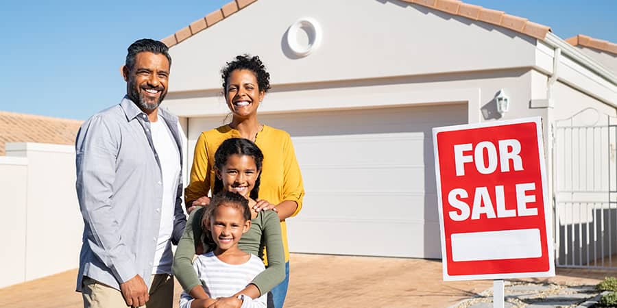 A family of four standing outside of their home next to a red for sale sign.
