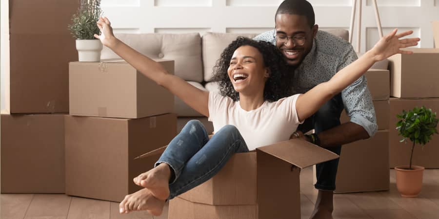 A woman sitting in a cardboard box with a man behind her pushing it across a living room.