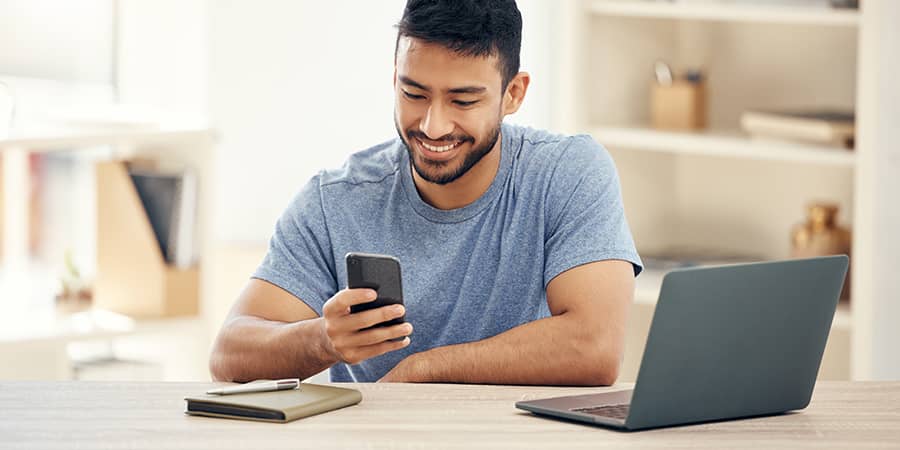 A young man sitting at a table with a laptop open and looking at a smartphone.