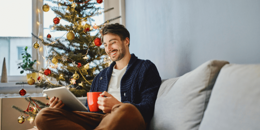A man sitting on a couch looking at a tablet with a christmas tree in the background.