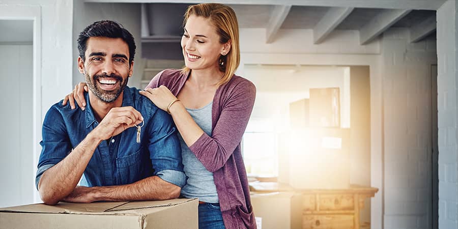 A man holding keys standing with a woman in a living room surrounded by cardboard boxes.