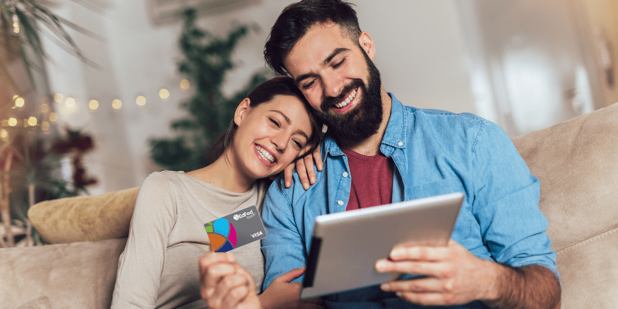 A man and woman siting on a couch while the man uses a tablet and holds the EdFed Rewards Visa Credit Card.