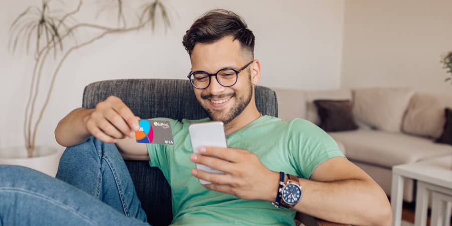 A young man sitting on a couch and holding the EdFed Rewards Visa credit card.