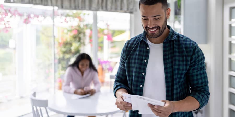 A man standing in a kitchen holding paper statements. In the background, a woman sitting at a table looking at documents.