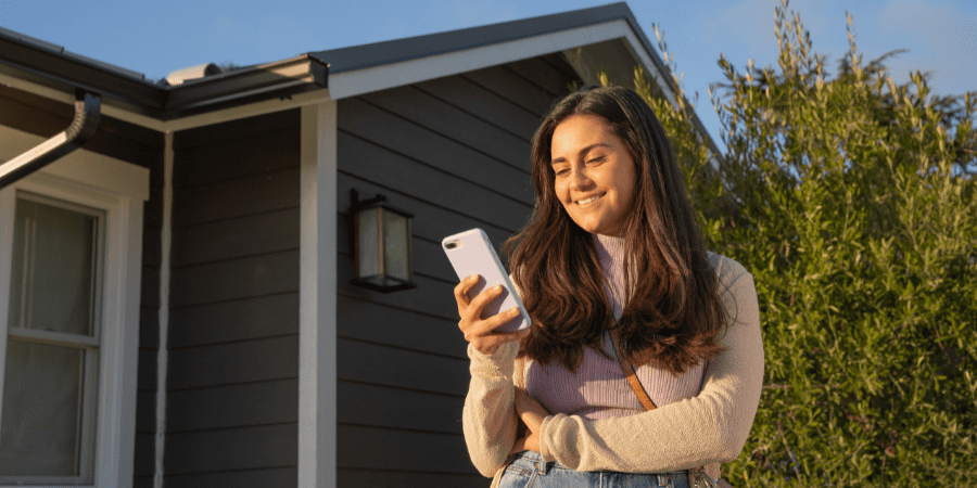 A young woman holding a smartphone standing outside a home.
