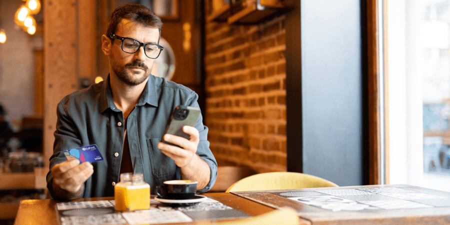 A man sitting at a coffee shop entering a credit card into his smartphone.