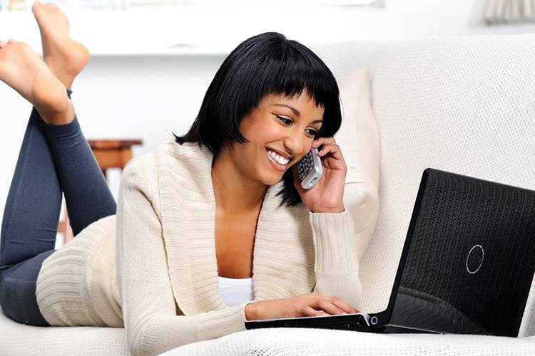 Woman laying on couch on portable phone and an open laptop in front of her.