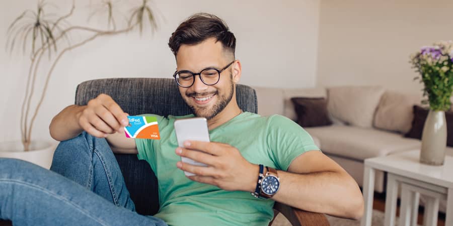 A young man sitting on a couch and holding the EdFed Rewards Visa credit card.