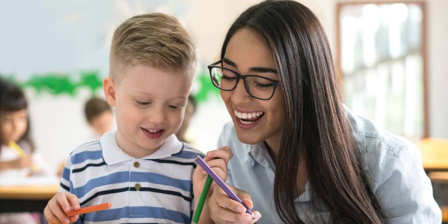A school teacher standing in front of a classroom with arms crossed and a big smile.