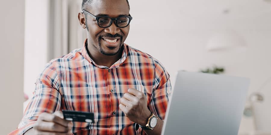 A young male excitedly sitting at a table using a laptop while holding a credit card.