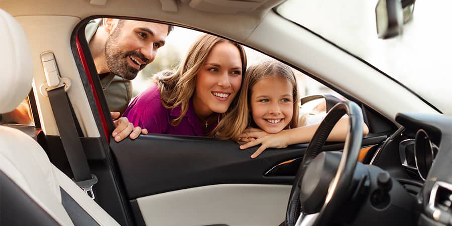 A smiling family looking through the driver's side window of a car.