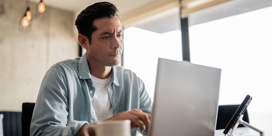 A man sitting at a desk working on a laptop with a concerned expression.