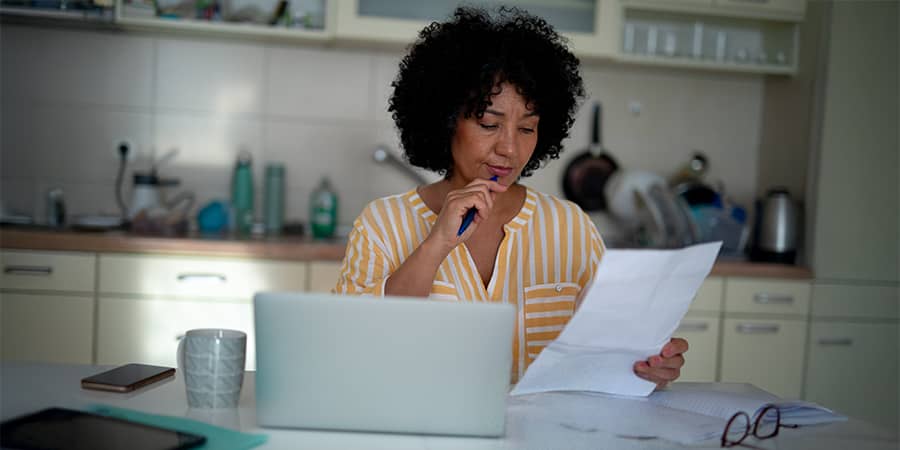 A woman sitting in her kitchen working on a laptop with a concerned expression.